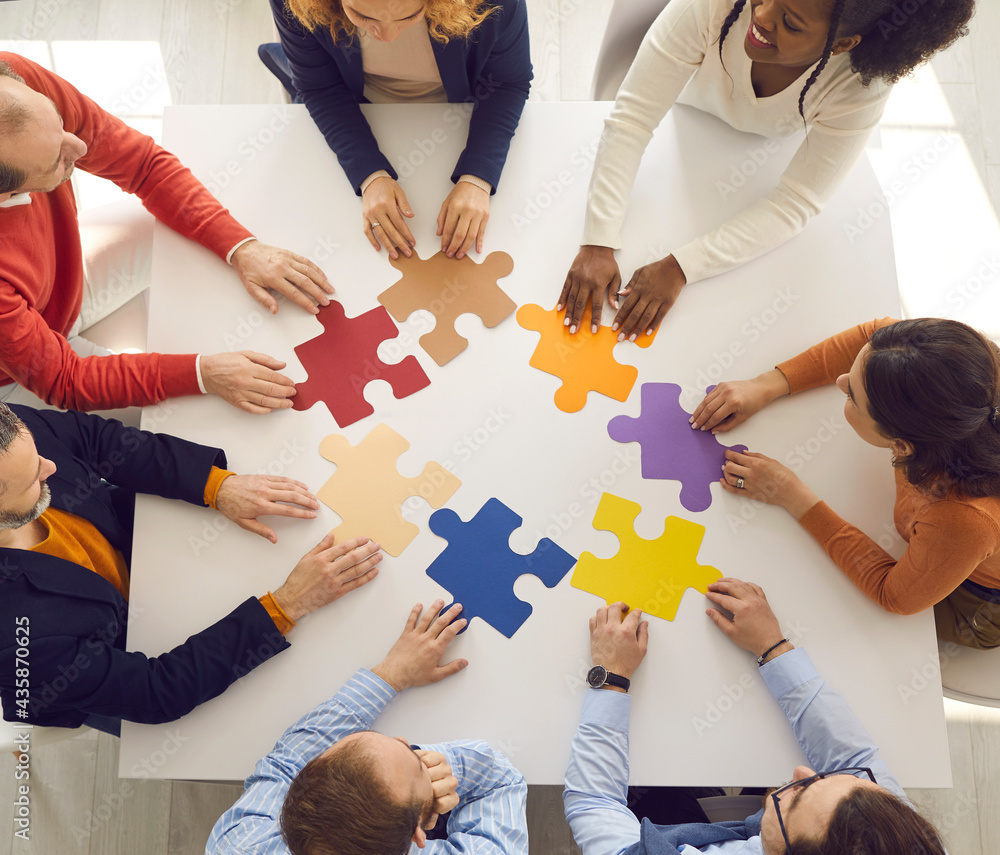 Team of young and senior employees sitting in circle around office table  putting jigsaw puzzle together as metaphor for teamwork and looking for  working business solutions. Square format, high angle Stock Photo
