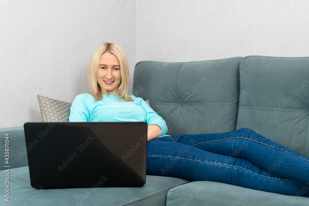 Young beautiful blonde woman with a smile uses a laptop while lying on the couch at home Young beautiful blonde woman with a smile uses a laptop while lying on the couch at home