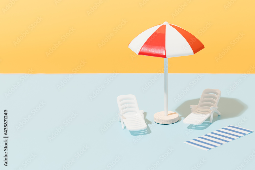 White and red beach umbrella, sun chairs and blue and white striped towel on blue and yellow background. Beach set for sunny days. Summer holiday concept. Copy space.