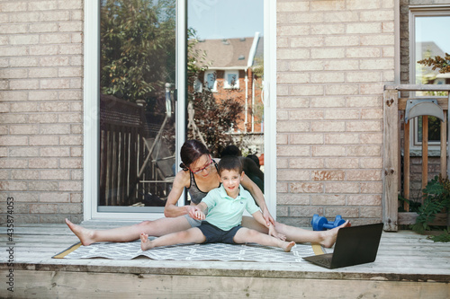 Family sport activity. Middle age Caucasian mother with toddler son doing distant remote online fitness workout on house backyard with laptop. Parent exercising with child boy kid. Sport at home.