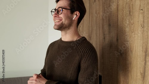 A smiling man in a sweater talking on wireless headphones and actively gesticulating while walking in the room