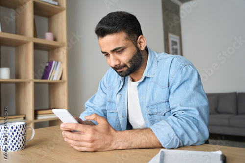 Young Indian Hispanic businessman using smartphone working remotely reading ebook from modern home office. Latin student having virtual training on cellphone using online tech applications.