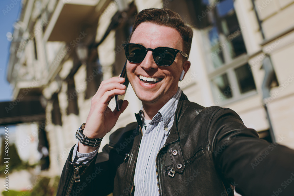 Close up young friendly fun happy caucasian man 20s wearing black leather jacket glasses doing selfie shot on mobile cell phone camera, talk outdoors Concept of people urban lifestyle spring season.