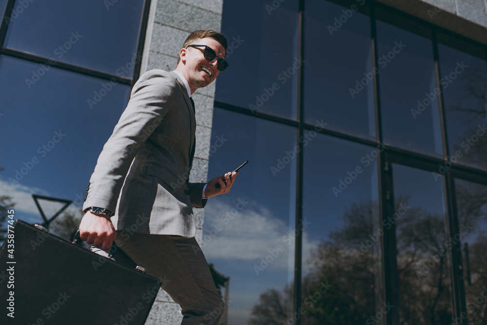 Bottom view young successful smiling employee business man 20s wear grey suit walking go near office glass wall building outdoors in downtown city talk hold mobile cell phone briefcase diplomat bag. Bottom view young successful smiling employee business man 20s wear grey suit walking go near office glass wall building outdoors in downtown city talk hold mobile cell phone briefcase diplomat bag.