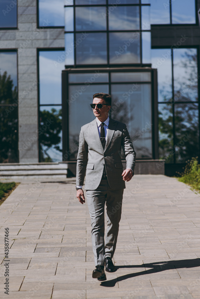Full length young confident serious employee business man 20s wearing grey suit walking go near office glass wall building outdoors in downtown city center look camera. Achievement career concept Full length young confident serious employee business man 20s wearing grey suit walking go near office glass wall building outdoors in downtown city center look camera. Achievement career concept