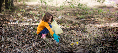 Caucasian kid girl cleaning up in the park. Child collect trash in the forest. Girl in a yellow turtleneck and jeans in a spring forest