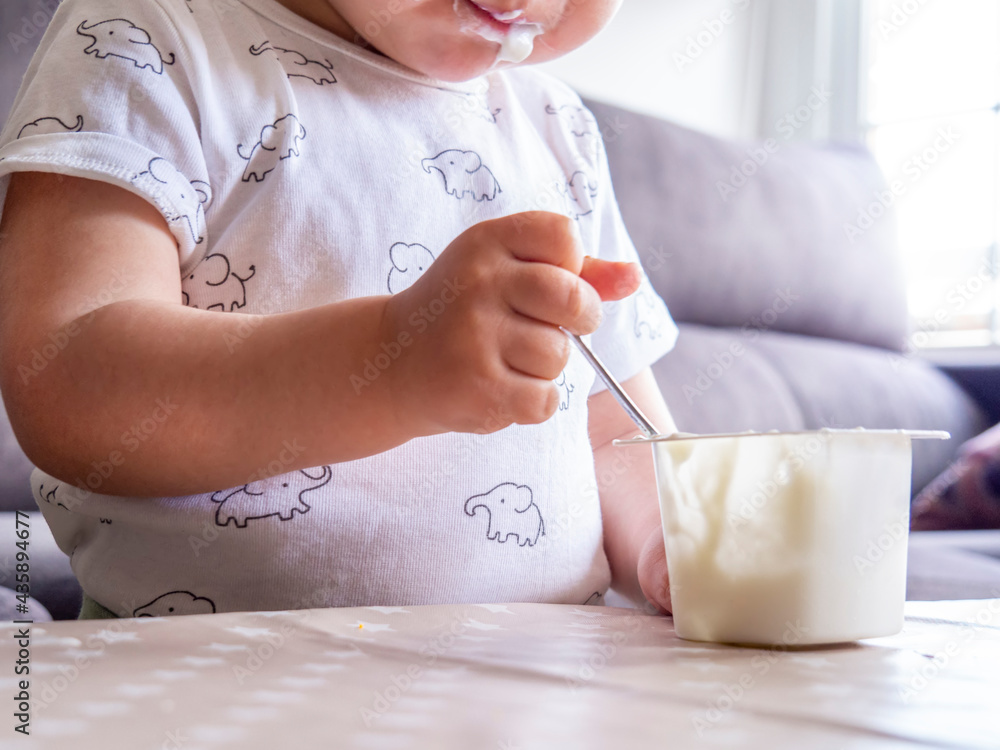 Baby boy hand holding a spoon inside a yogurt while eating. Baby learning how to eat, domestic life with a toddler. Unrecognizable baby with dirty mouth while eating delicious food. Baby boy hand holding a spoon inside a yogurt while eating. Baby learning how to eat, domestic life with a toddler. Unrecognizable baby with dirty mouth while eating delicious food.
