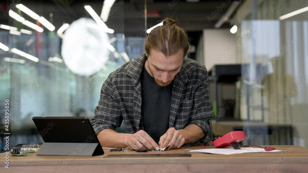 Young Man In The Workshop Is Engaged In Soldering. Chips And Devices On The Table. Young Man In The Workshop Is Engaged In Soldering. Chips And Devices On The Table.