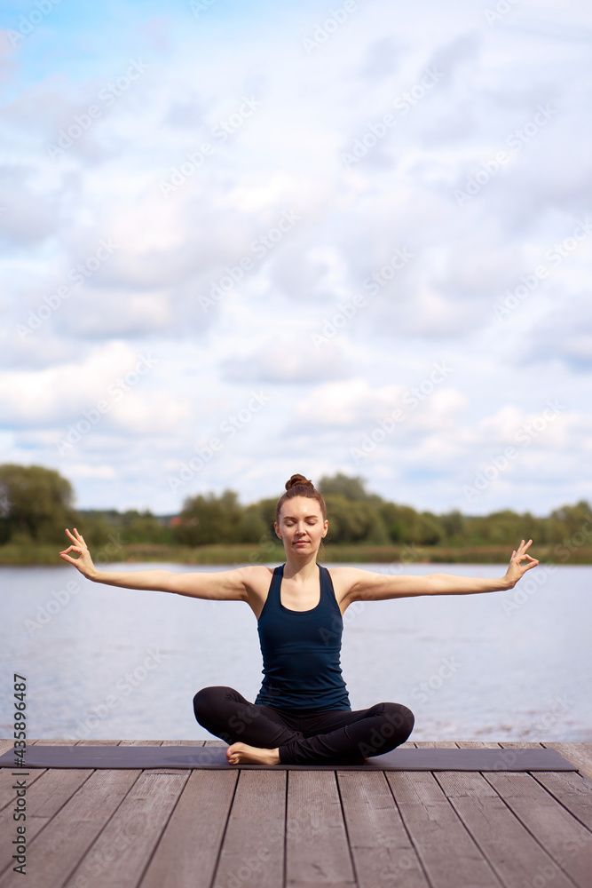Young attractive woman with closed eyes practicing yoga on the wooden pier near lake. Outdoor activities.