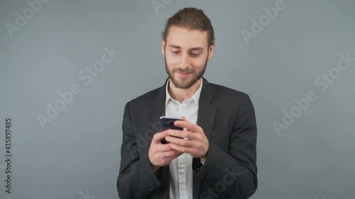 Young man businessman in a business suit pulls a smartphone from his jacket pocket and reads a message from his beloved. The man gently smiles. Man isolated on gray background in studio. 4K