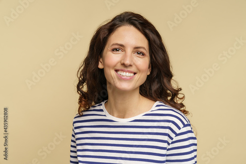 Portrait of curly caucasian female, closeup studio shot over beige background. Millennial woman with happy toothy smile and clean skin dressed in stripped t-shirt. Natural beauty and no makeup concept