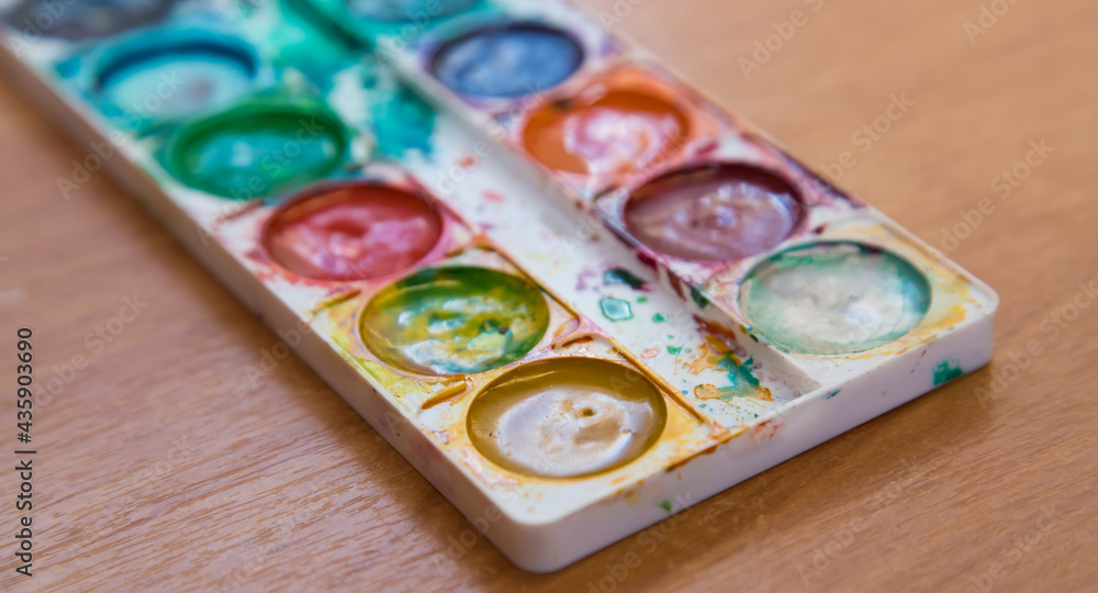 On the table is a used set of multicolored watercolor paints for painting. Top view of half-empty jars of watercolors. Flat layout on the table. On the table is a used set of multicolored watercolor paints for painting. Top view of half-empty jars of watercolors. Flat layout on the table.