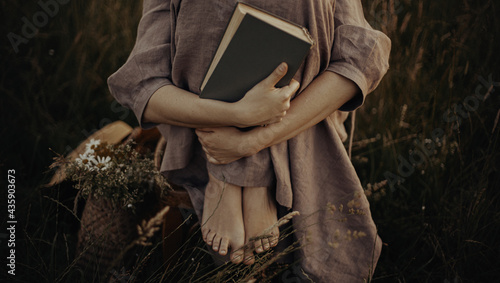 Beautiful woman in linen dress sitting barefoot on rustic chair with book in summer meadow, close up. Young female holding book and relaxing in countryside. Atmospheric stylish vintage image