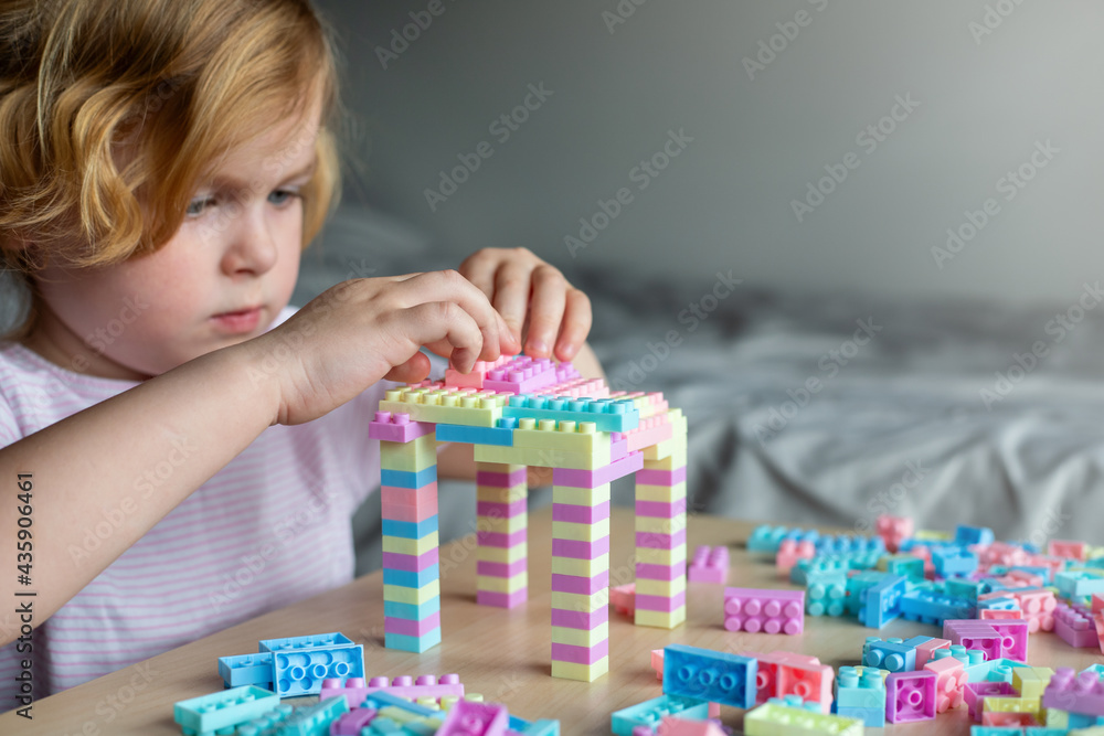 Fototapeta premium Small cute preschooler girl playing with colorful toy building blocks, sitting at the table.