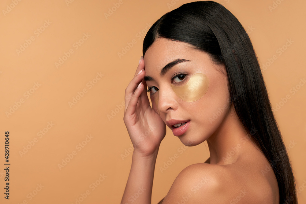 Young Asian woman in white lingerie and clean radiant skin with moisturizing patches under the eyes on a beige background. Spa care, cosmetology. Young Asian woman in white lingerie and clean radiant skin with moisturizing patches under the eyes on a beige background. Spa care, cosmetology.