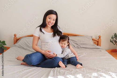 Asian Chinese pregnant woman with toddler girl sitting on bed at home. Girl daughter kid hugging mom belly. Mother and baby daughter expecting waiting for a new family member. Ethnic diversity