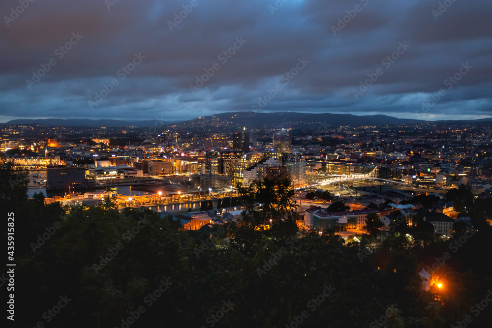 Night panoramic view of Oslo, capital of Norway. Dark cloudscape ower capital of Norway. Scandinavian city with lighted buildings and bridges, surrounded by forests.