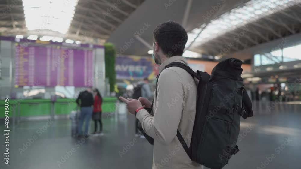 Masked man with backpack happy to travel again, using smart phone near flight information board. Male traveler in protective mask watching and waiting for flight schedule on boarding time monitor Masked man with backpack happy to travel again, using smart phone near flight information board. Male traveler in protective mask watching and waiting for flight schedule on boarding time monitor
