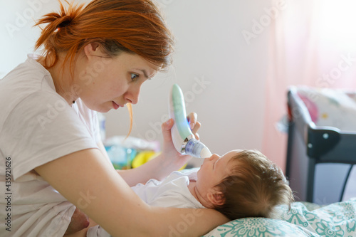 Side view of caucasian woman mother using electric baby nasal aspirator mucus nose suction sucking the saliva from baby s nose cleaning while lying on the bed at home in day motherhood care concept