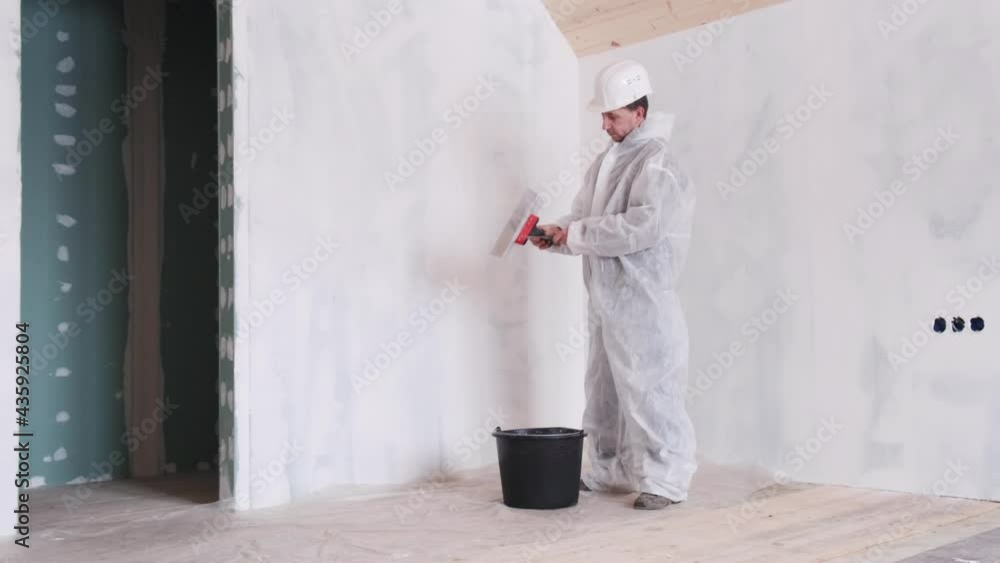 A full-length young man with a spatula in his hands and a protective suit makes repairs in the house. Leveling the walls with plaster. The concept of renovation in the house. A full-length young man with a spatula in his hands and a protective suit makes repairs in the house. Leveling the walls with plaster. The concept of renovation in the house.
