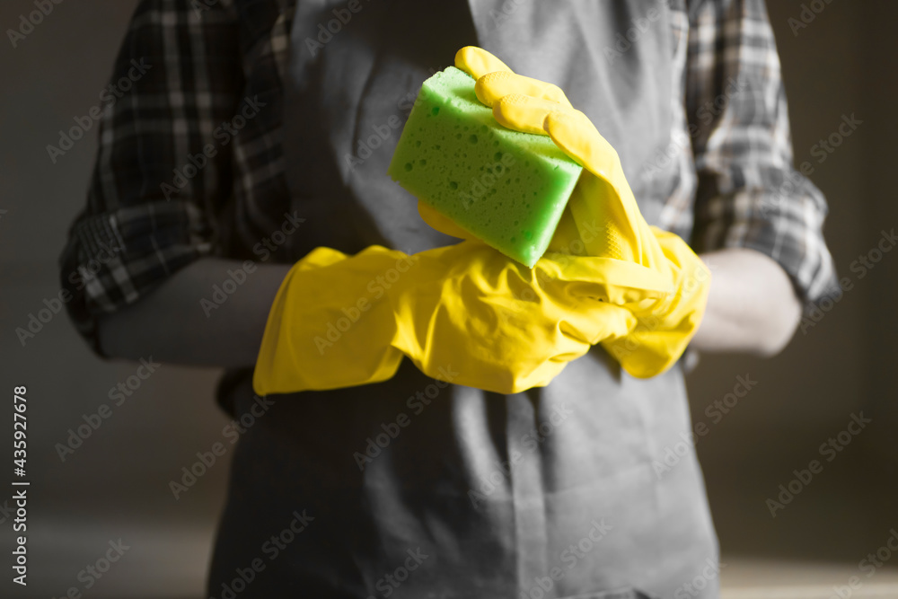 Young female hands in yellow rubber gloves hold a new green sponge for cleaning house and wiping surfaces. A female housekeeper in a plaid shirt and a gray apron holds a rag and plans to disinfect. Young female hands in yellow rubber gloves hold a new green sponge for cleaning house and wiping surfaces. A female housekeeper in a plaid shirt and a gray apron holds a rag and plans to disinfect.