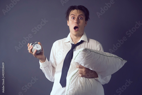 A young funny man in a white shirt and tie with disheveled hair slept through the deadline, holding a vintage alarm clock in his hand. The office worker is overworked and tired.