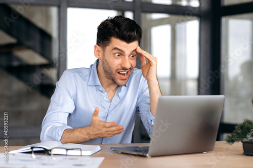 Caucasian young man, manager, freelancer or office worker, sitting at table in office, using laptop, shocked by news or message, looking at screen in surprise, gesturing with his hand, puzzled