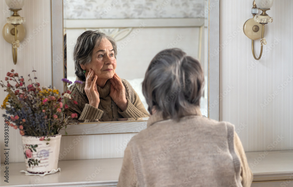 An elderly well-groomed gray-haired woman aged 80+ looks at herself in the mirror while sitting at the cosmetic table. Old lady, pensioner does facial skin care in her cozy home. An elderly well-groomed gray-haired woman aged 80+ looks at herself in the mirror while sitting at the cosmetic table. Old lady, pensioner does facial skin care in her cozy home.