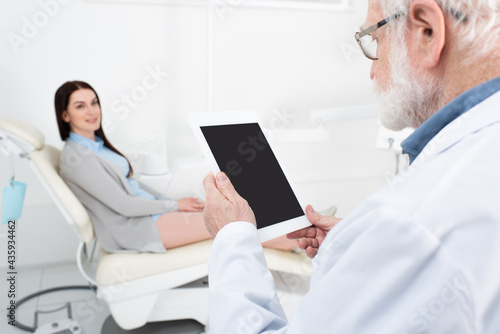senior dentist giving consultation to blurred patient in dental chair and holding tablet with blank screen in clinic.
