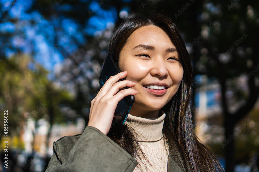 young multiracial Asian-American businesswoman, laughing while talking on the phone with a smart phone, outside in a city park.
