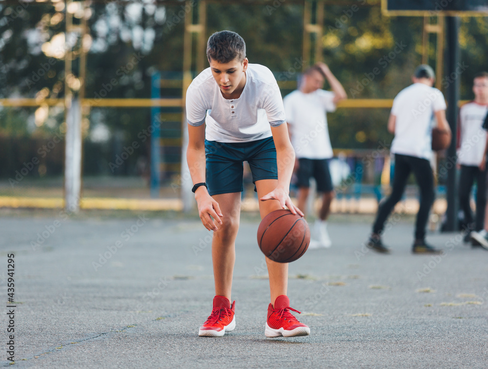 Cute boy in white t shirt plays basketball on a city playground. Active teen enjoying outdoor game with orange ball. Hobby, active lifestyle, sport for kids.	
