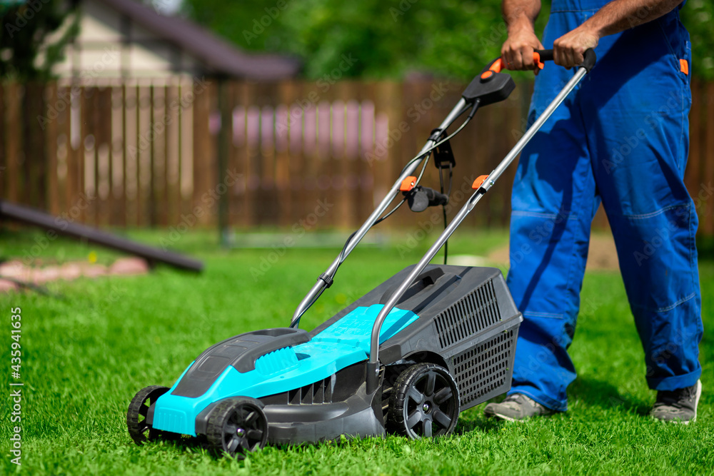 Close-up of a man in overalls with a lawn mower cutting green grass in a modern garden. Lawn mowing machine. Close-up of a man in overalls with a lawn mower cutting green grass in a modern garden. Lawn mowing machine.