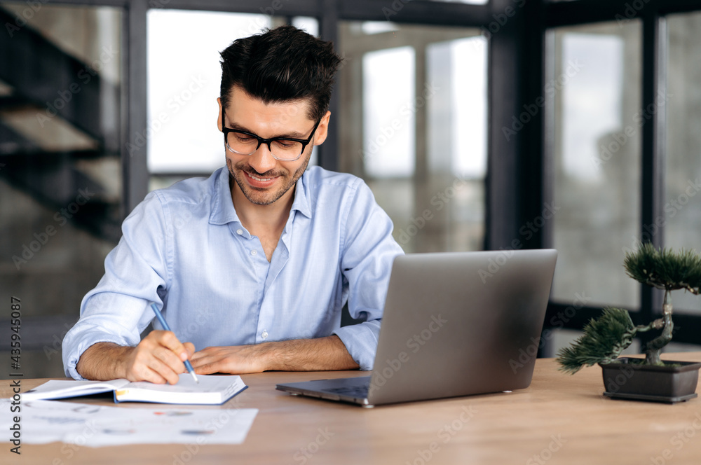 Fototapeta premium Concentrated satisfied successful caucasian businessman, freelancer or broker wearing glasses, sitting at a table in a modern office, using a laptop, taking notes, taking online training, smiling