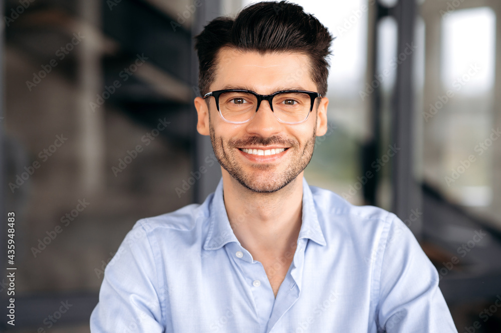 Close up portrait of handsome confident successful young caucasian man wearing glasses, wearing stylish shirt, sitting at office, looking directly at camera and smiling friendly Close up portrait of handsome confident successful young caucasian man wearing glasses, wearing stylish shirt, sitting at office, looking directly at camera and smiling friendly
