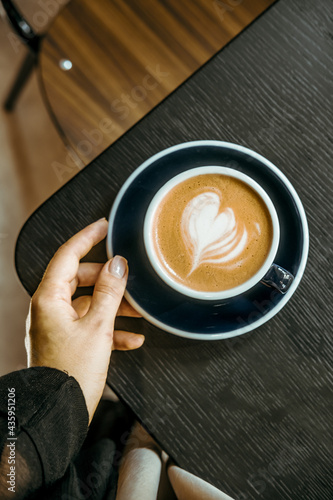 Woman's hand holding cappuccino coffee on black wooden table at coffee shop. Food drink photography concept. Lovely, calm, tasty, atmospheric moody shot. Close up, top view