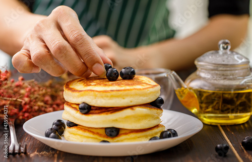 Close up hand of unrecognizable woman holding hand decorating Pancake by picking up blueberries and blur teapot background. Concept sweet food.