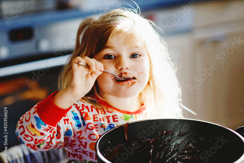 Foto Cute toddler girl eating chocolate dough rests with spoon and fingers from pot