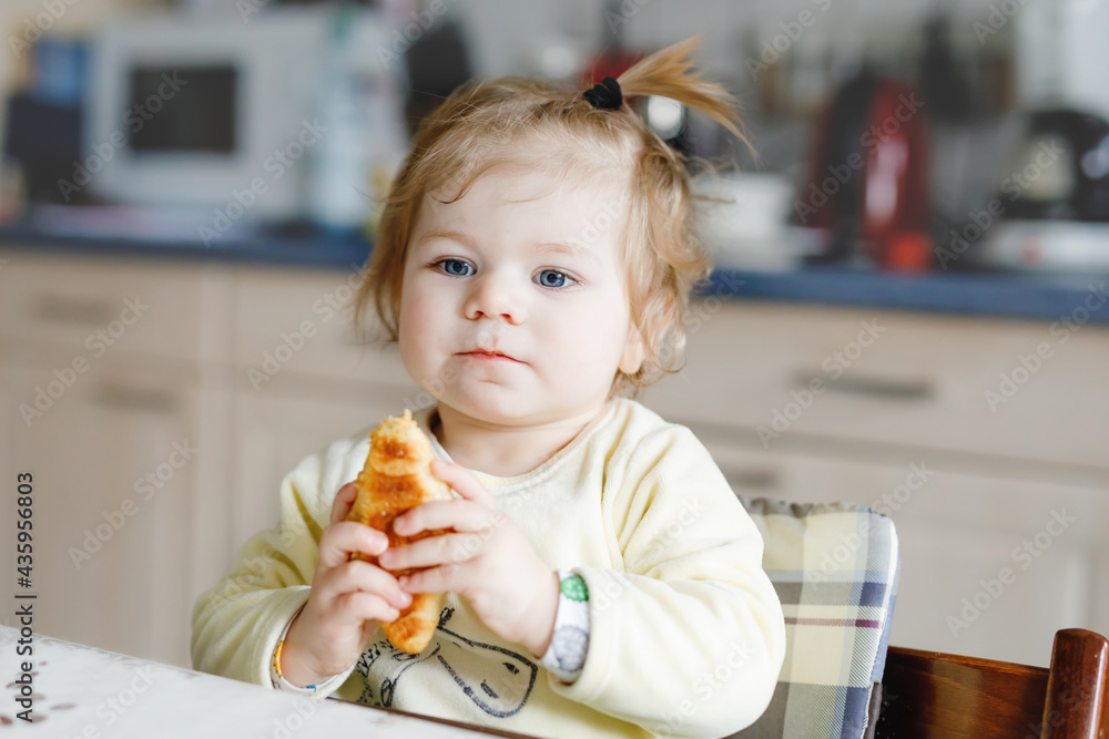 Happy little baby girl eating fresh croissant for breakfast or lunch. Healthy eating for children. Toddler child in colorful pajama sitting in domestic kitchen after sleeping in the morning, Happy little baby girl eating fresh croissant for breakfast or lunch. Healthy eating for children. Toddler child in colorful pajama sitting in domestic kitchen after sleeping in the morning,