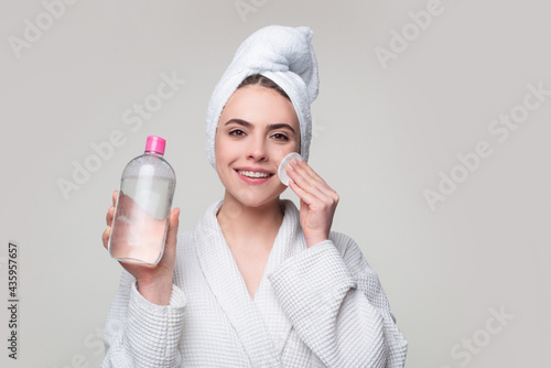 Girl with micellar water. Beautiful woman with towel on head, micellar water and cotton pad isolated on gray. photo