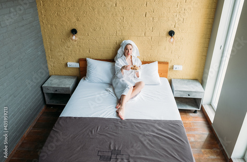 A woman in a white robe at the hotel has breakfast on the bed. 