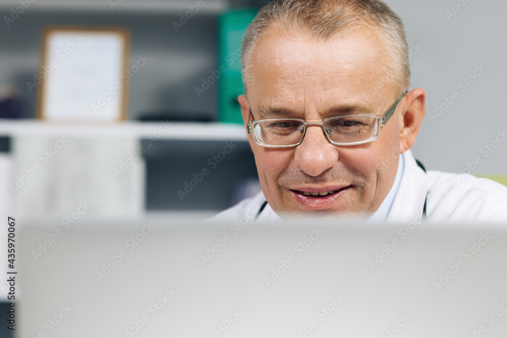 Physician in White Lab Coat is Browsing Medical History Behind a Desk in Hospital Office. Family Medical Doctor in Glasses is Working on a Laptop Computer in a Health Clinic Physician in White Lab Coat is Browsing Medical History Behind a Desk in Hospital Office. Family Medical Doctor in Glasses is Working on a Laptop Computer in a Health Clinic