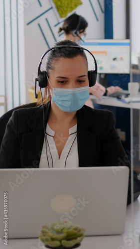 Freelancer with protective face mask wearing headset while talking into microphone about business meeting. Businesswoman working on laptop computer in company office during coronavirus pandemic