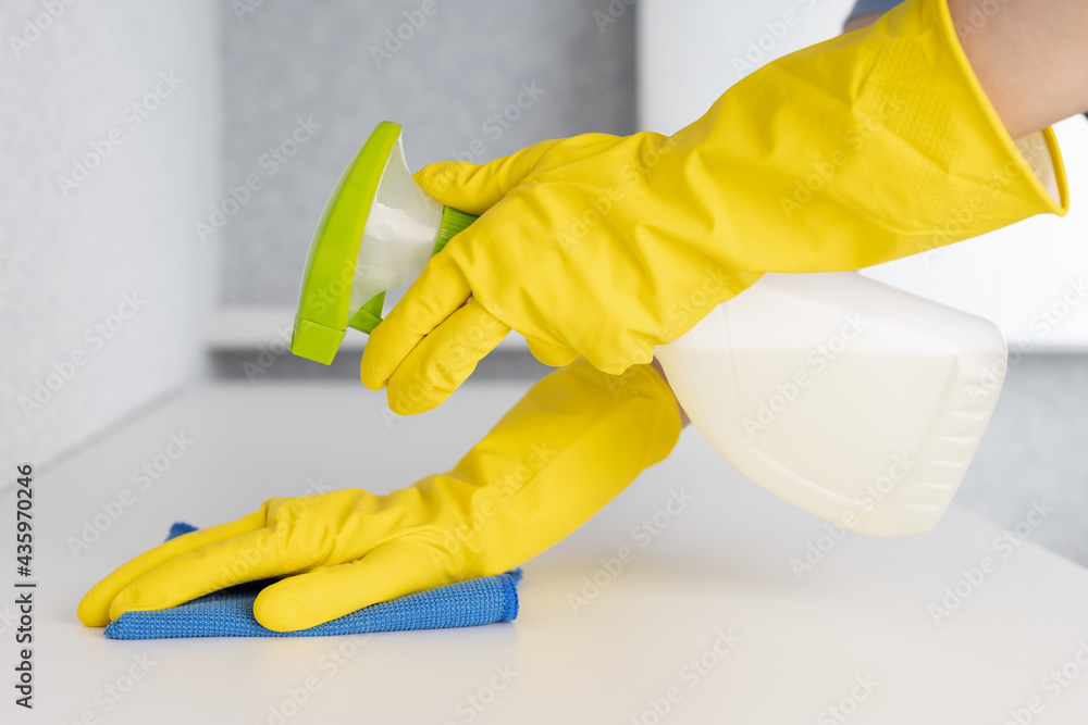 Woman in yellow rubber gloves cleaning table with blue cloth and spray.