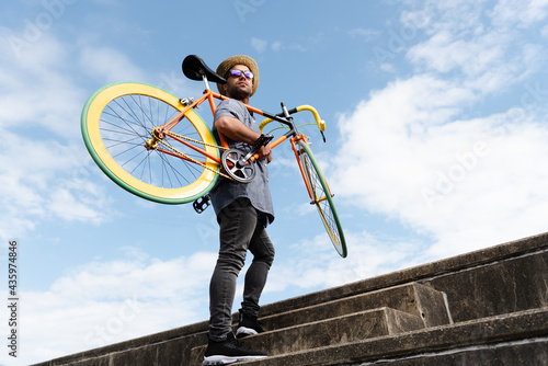 Hispanic hipster carrying a colorful fixie bicycle on his shoulder while climbing the stairs.