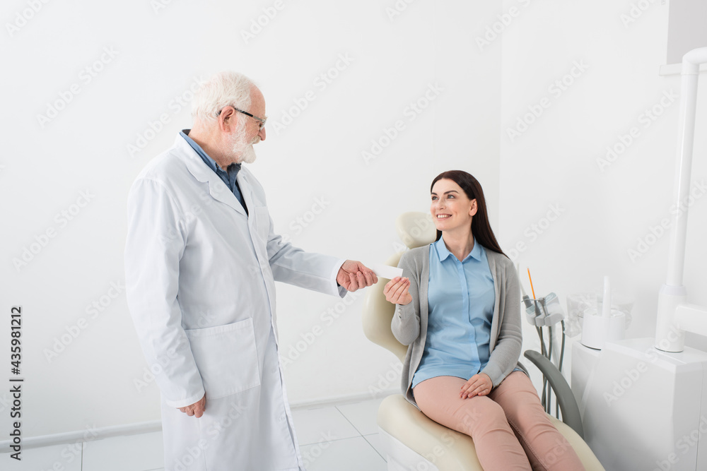 Obraz premium cheerful senior dentist giving empty card to smiling woman in dental chair in clinic.