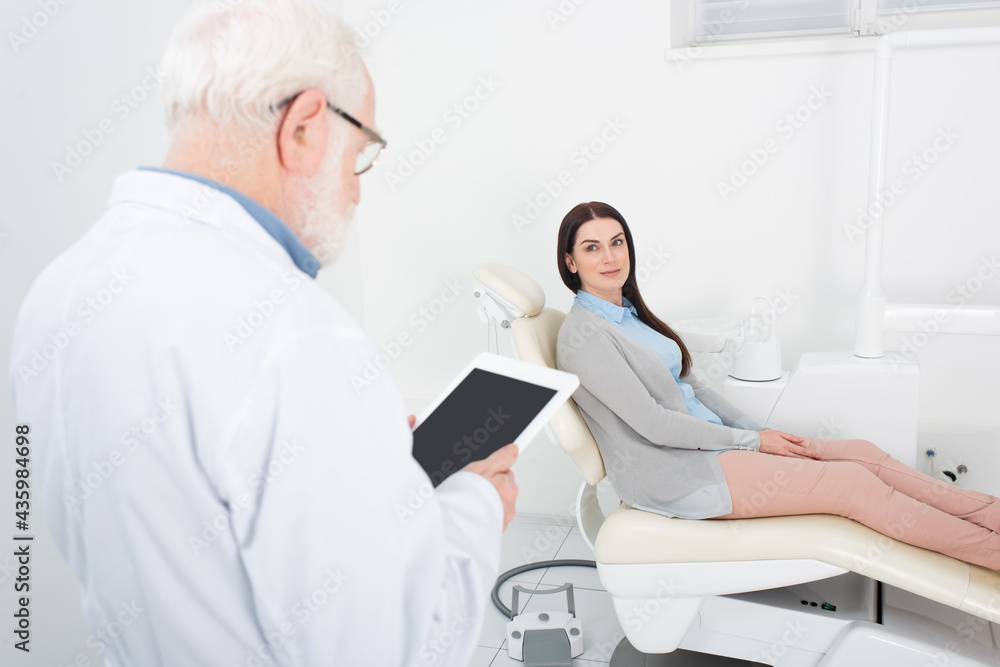 senior dentist looking at blank screen tablet in front of blurred patient in dental chair in clinic. senior dentist looking at blank screen tablet in front of blurred patient in dental chair in clinic.