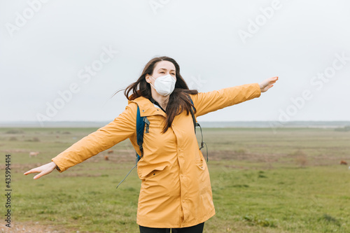 Woman with mask outside in the dune landscape. Tourist in a yellow rain jacket holds her arms wide open as if she is flying. Wearing mask outdoors. Vacation on the North Sea during the coronavirus 