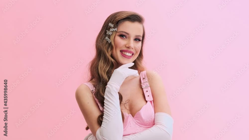 A happy young woman wearing gorgeous evening pink dress with gloves and hairpins is smiling to the camera standing isolated over a pink wall in the studio A happy young woman wearing gorgeous evening pink dress with gloves and hairpins is smiling to the camera standing isolated over a pink wall in the studio
