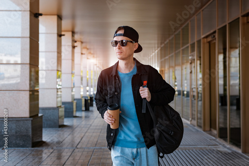 young hipster guy stands city street black cap, holding cup coffee. background modern street glass corridor