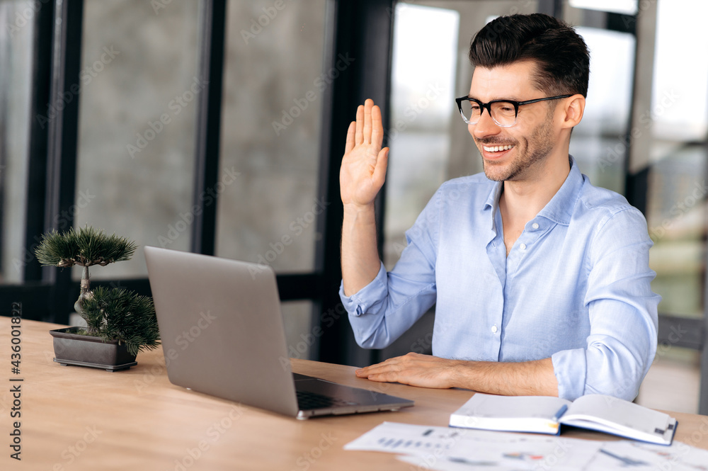 Fototapeta premium Cute friendly young Caucasian businessman, manager or freelancer wearing glasses, sitting at a laptop, communicating via video conference with friends or colleagues, gesturing with his hand, smiling